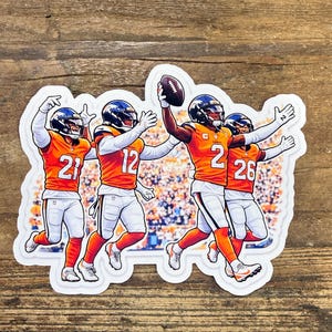 May include: A sticker featuring four football players in orange and white uniforms, with helmets. One player holds a football. The players are in action, with arms raised. The background shows a blurred crowd.