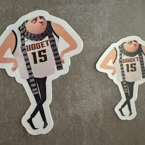 May include: Two stickers of a cartoon character wearing a white tank top with the word "UGGET" and the number "15" on it. The character is wearing a black and white striped scarf and black pants. The stickers are on a gray background.