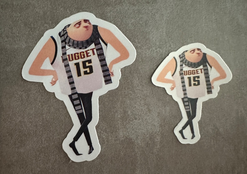 May include: Two stickers of a cartoon character wearing a white tank top with the word "UGGET" and the number "15" on it. The character is wearing a black and white striped scarf and black pants. The stickers are on a gray background.