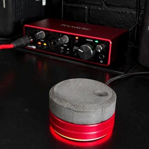 Può includere: Una manopola cilindrica per il controllo del volume con una parte superiore in cemento grigio testurizzato e una base in metallo rosso liscio con un anello luminoso a LED rosso integrato sul fondo. Il dispositivo è davanti a un'interfaccia audio Focusrite rossa.
