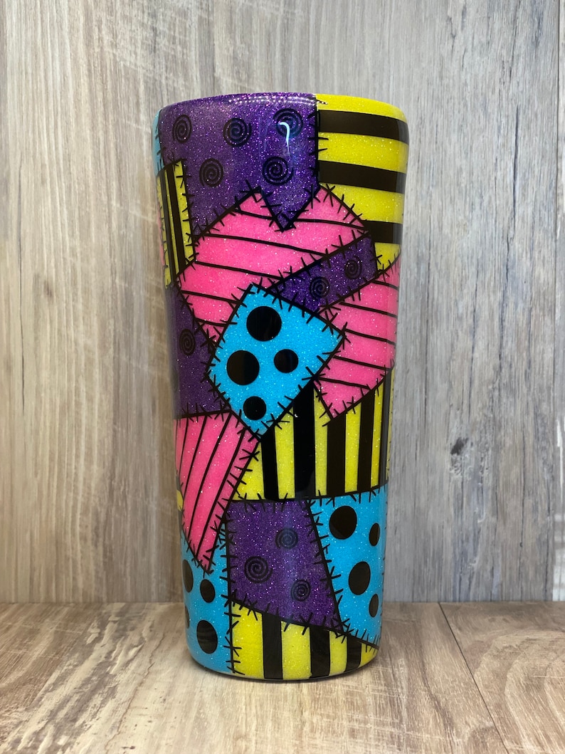 May include: A tall, stainless steel tumbler with a patchwork design featuring various colors, including pink, blue, yellow, and purple. The design includes black polka dots and stripes, and the tumbler has a glitter finish.