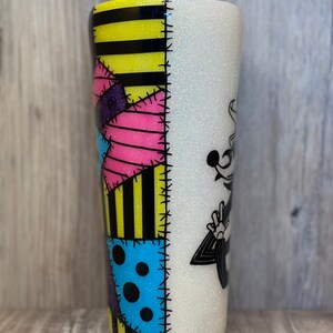 May include: A white glitter tumbler with a black and yellow striped design on one side and a black and white illustration of Jack Skellington from The Nightmare Before Christmas on the other side. The design on the striped side is a patchwork of different colored fabrics with stitching details.