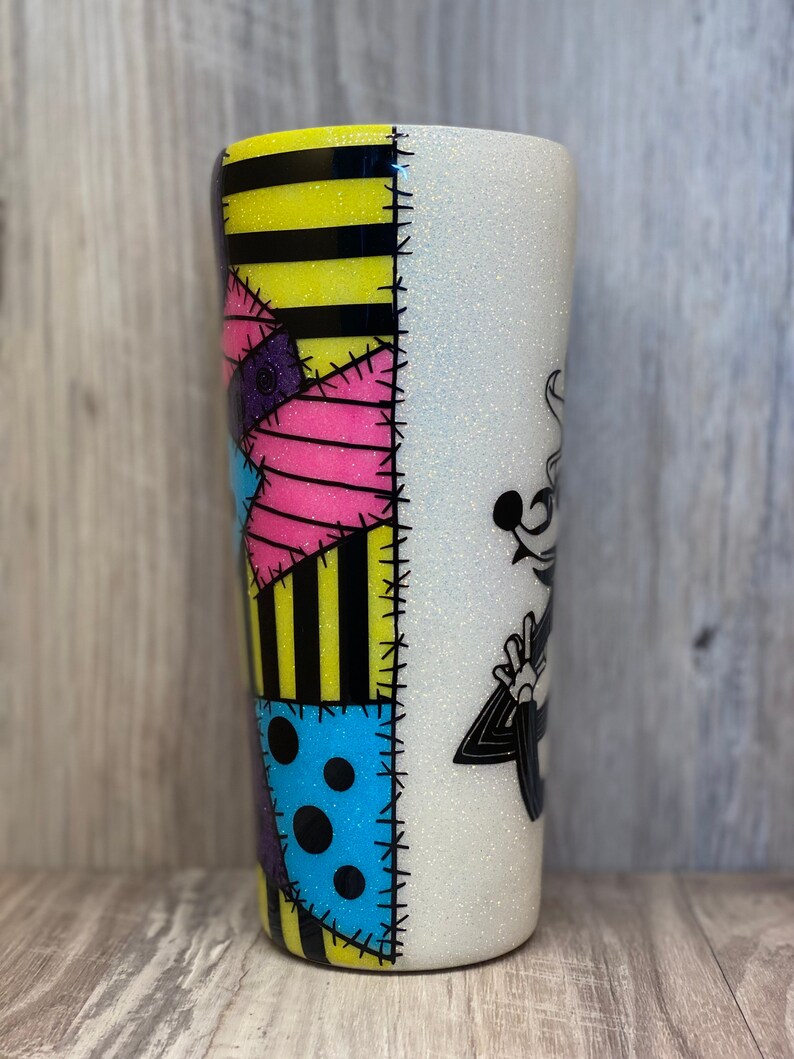 May include: A white glitter tumbler with a black and yellow striped design on one side and a black and white illustration of Jack Skellington from The Nightmare Before Christmas on the other side. The design on the striped side is a patchwork of different colored fabrics with stitching details.