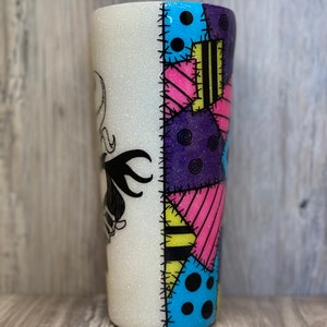 May include: A white glitter tumbler with a black and white image of Sally from the Nightmare Before Christmas on one side. The other side features a colorful patchwork design with blue, yellow, pink, and purple.