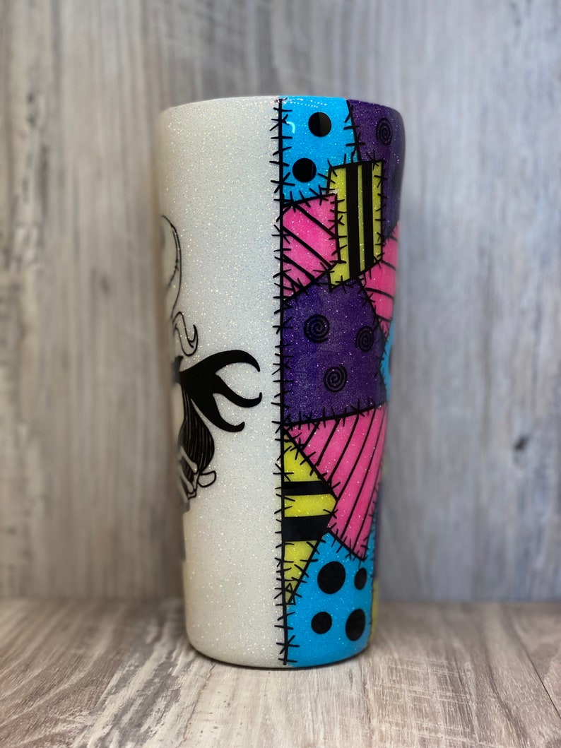 May include: A white glitter tumbler with a black and white image of Sally from the Nightmare Before Christmas on one side. The other side features a colorful patchwork design with blue, yellow, pink, and purple.