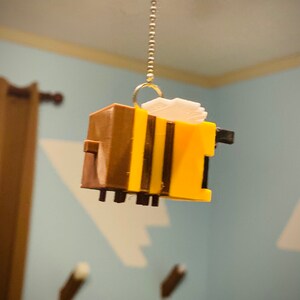 May include: A yellow and brown bee-shaped ornament hanging from a gold chain. The bee has black stripes and a white wing.
