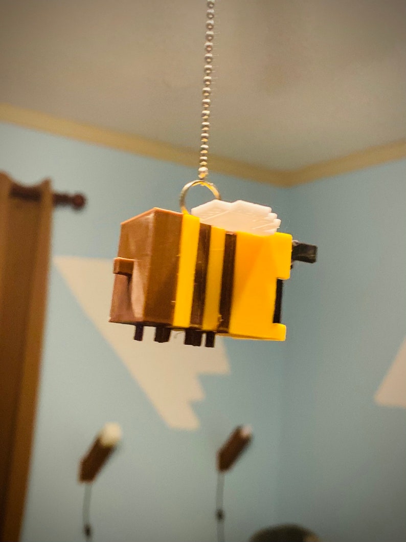 May include: A yellow and brown bee-shaped ornament hanging from a gold chain. The bee has black stripes and a white wing.