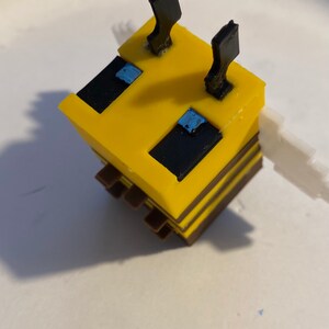 May include: A yellow and black bee-shaped toy with black antennae and blue eyes. The toy is made of plastic and has a blocky design.