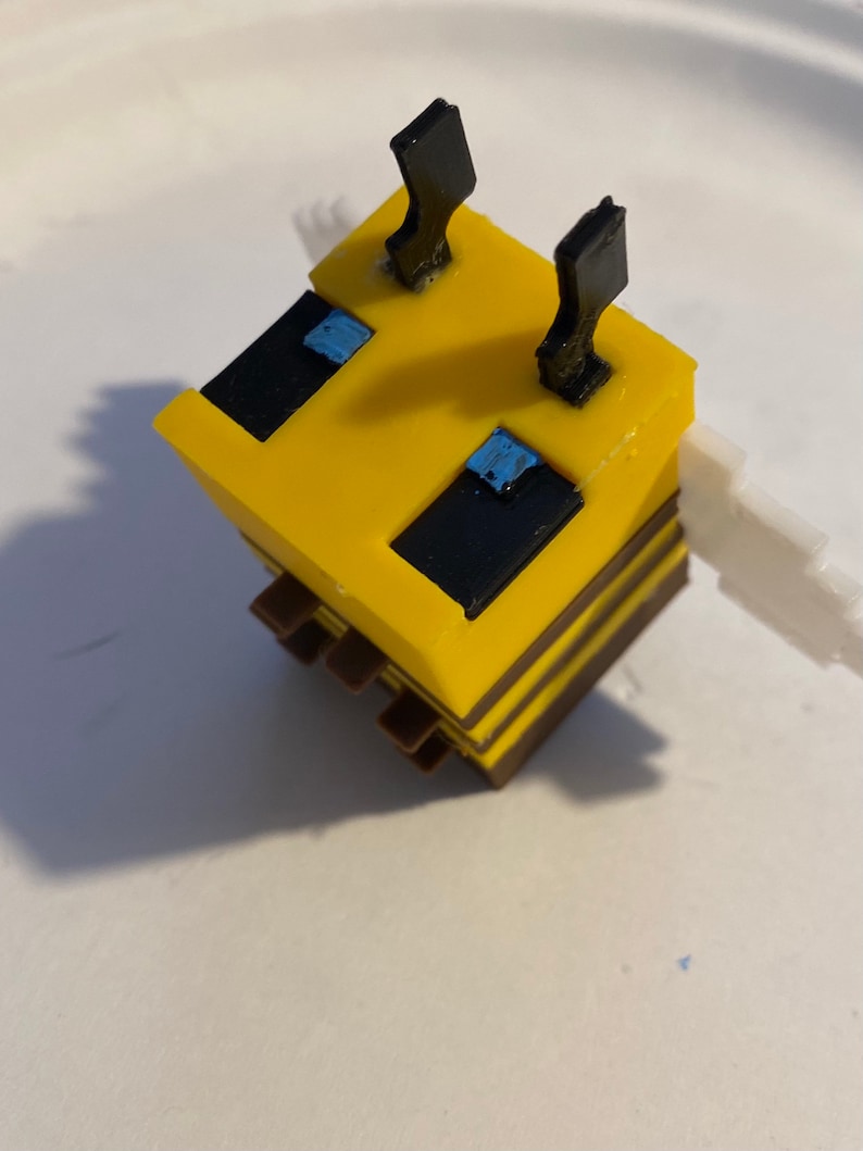 May include: A yellow and black bee-shaped toy with black antennae and blue eyes. The toy is made of plastic and has a blocky design.