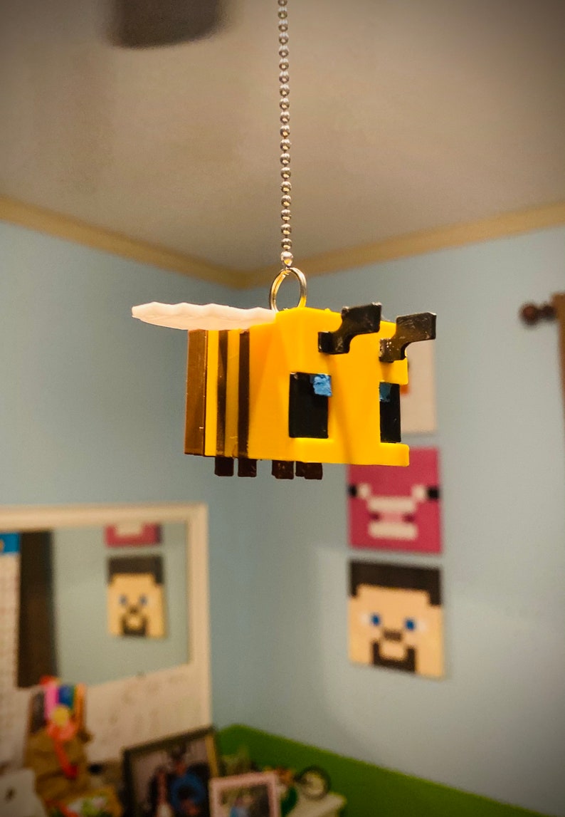 May include: A yellow and black Minecraft bee charm hanging from a silver chain. The bee has a white wing and blue eyes.