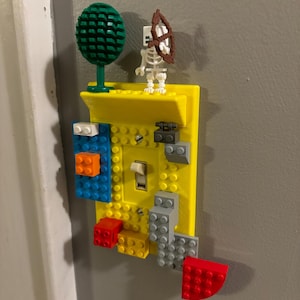 Könnte beinhalten: Ein gelber Lichtschalter-Abdeckung, die mit bunten Lego-Steinen verziert ist, darunter ein grüner Baum, eine Skelettfigur mit Pfeil und Bogen und eine Vielzahl von roten, blauen und grauen Steinen.