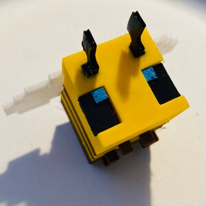 May include: A yellow and black bee-shaped Minecraft figure with blue eyes and white wings. The figure is made of plastic and has a blocky design.