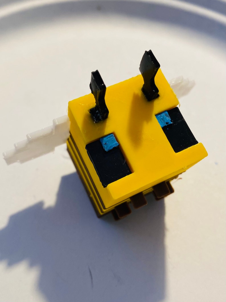 May include: A yellow and black bee-shaped Minecraft figure with blue eyes and white wings. The figure is made of plastic and has a blocky design.
