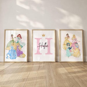 May include: Three framed art prints with watercolour illustrations of Disney princesses. The prints feature Cinderella, Ariel, Belle, and Rapunzel. The central print has a pink letter "H", the name "Hollie" and a gold crown.