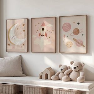 Op de afbeelding: Drie ingelijste kunstprints met een ruimtethema. De prints tonen een maan, een raket en planeten. Het kleurenpalet omvat roze, beige en goud. Twee teddyberen en een houten huis staan op een bank onder de prints.
