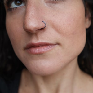 May include: Close-up of a person's face with a dark nose ring. The nose ring has a wavy design. The person has dark hair and is looking up. The skin tone is light with a natural makeup look.