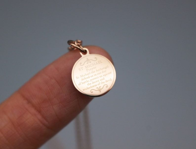 May include: A gold-toned pendant necklace with a round charm featuring an engraved prayer to Saint Michael the Archangel. The charm has a decorative border and the text reads "St. Michael the Archangel, defend us in our day of battle, protect us against the deceit and wickedness of the devil."