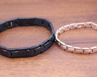 May include: Two magnetic bracelets, one black and one rose gold, on a wooden surface.