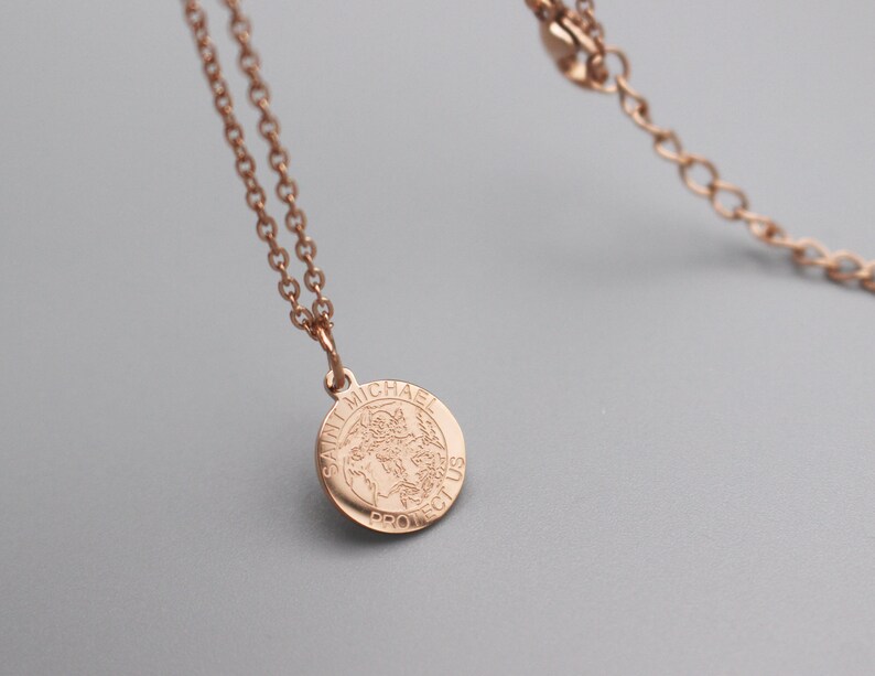 May include: A rose gold chain necklace with a round pendant featuring the words "Saint Michael Protect Us".