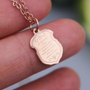 May include: A rose gold necklace with a shield-shaped pendant that has the St. Michael prayer engraved on it. The prayer reads: "St. Michael the Archangel, defend us in the day of battle. Be our protection against the wickedness of the enemy."