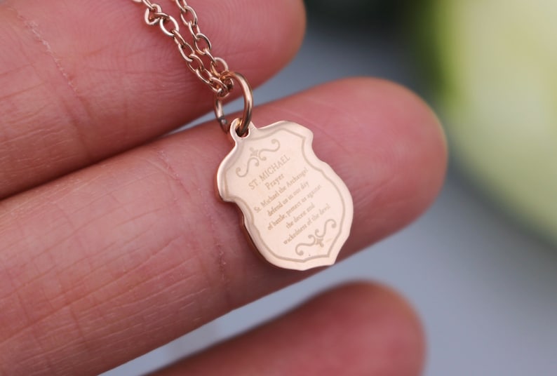 May include: A rose gold necklace with a shield-shaped pendant that has the St. Michael prayer engraved on it. The prayer reads: "St. Michael the Archangel, defend us in the day of battle. Be our protection against the wickedness of the enemy."