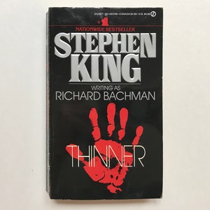 May include: A black paperback book with a red handprint on the cover. The title "THINNER" is written in white letters. The author is Stephen King, writing as Richard Bachman. The book is a nationwide bestseller.