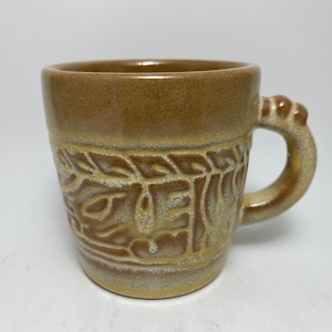 以下が含まれることがあります： 白く釉薬がかけられた茶色の陶器製のマグカップ。葉と蔓のレリーフ模様が施されています。マグカップには丸いノブが付いた取っ手があります。