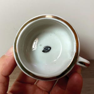 May include: White ceramic teacup with a gold rim and handle. The interior features a black and white eye design. The cup is held in a person's hand.