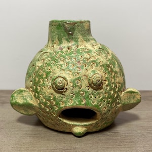 May include: A whimsical, green ceramic sculpture of a fish-like creature with large, round eyes, a wide open mouth, and small fins. The sculpture has a textured surface with a distressed, aged appearance, and a small opening at the top.