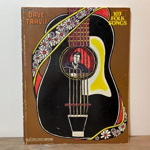May include: A vintage music book titled "Dave Travis Easy Guitar 103 Folk Songs." The cover features a black guitar illustration with a portrait of a man inside. The guitar is decorated with red, yellow, and white floral accents, set against a brown background.