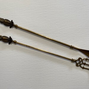May include: A set of two antique brass fireplace tools. One tool has a spoon-shaped end and the other has a three-pronged fork.