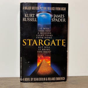 Peut inclure: Un livre de poche intitulé "Stargate" avec une couverture sombre représentant une pyramide et un portail bleu. La couverture comprend les noms de Kurt Russell et James Spader, et le texte "It will take you a million light years from home."