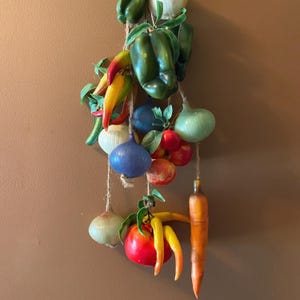 May include: A decorative wall hanging of artificial vegetables, including green peppers, red tomatoes, yellow peppers, and an orange carrot. The vegetables are strung together with twine, creating a rustic and colourful display.