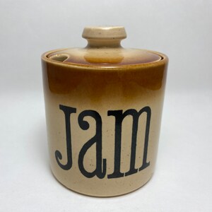 May include: A brown and beige ceramic jar with a lid. The jar has the word "Jam" written in black lettering.