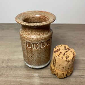 May include: A small, brown ceramic jar with the word "OREG" embossed on the side. The jar has a wide rim and a cork stopper. The jar and stopper have a rustic, handmade appearance.