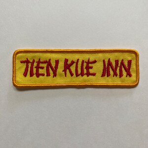Peut inclure: Écusson brodé jaune et orange avec le texte "Tien Kue Inn" en rouge.