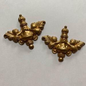 May include: Two gold-toned metal decorative embellishments with a floral design. Each embellishment features a central ornate element with a rounded top and a pointed bottom, surrounded by stylized leaves.