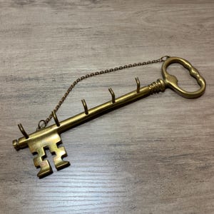 Puede incluir: Un colgador de pared con forma de llave de latón con un diseño decorativo. La llave presenta un asa, una serie de ganchos para colgar objetos y un paletón decorativo. Una cadena está unida a la llave para colgarla.