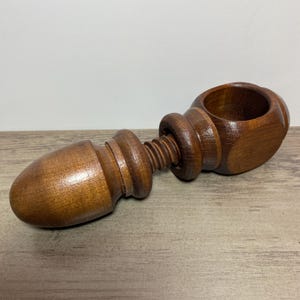 May include: A wooden nutcracker with a dark brown finish. The nutcracker has a bulbous end, a threaded central section, and a circular opening. The wood grain is visible, and the tool is lying on a wooden surface.