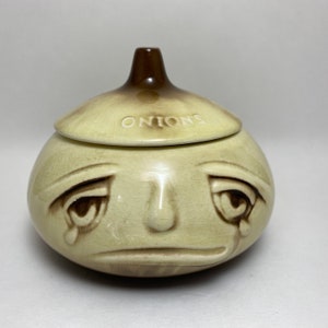 May include: A ceramic onion-shaped container with a crying face and the word "ONIONS" on the lid. The container is light brown and cream colored.