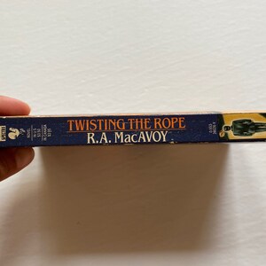 Puede incluir: Un libro de bolsillo azul con el t&iacute;tulo "Twisting the Rope" de R.A. MacAvoy. La portada del libro presenta la silueta de una persona con traje.