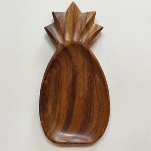 May include: A wooden pineapple-shaped serving tray. The tray is carved from a rich, dark brown wood, with the pineapple's crown and body clearly defined. The tray's surface has a smooth finish, showcasing the natural wood grain.