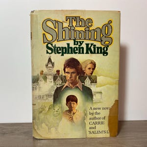 May include: Vintage paperback book cover for Stephen King's "The Shining." The cover features the title in large, bold letters with an illustration of a man, woman, and child. The cover also includes an illustration of a building and a dog.