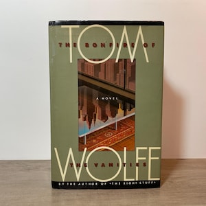 May include: A hardback book titled "The Bonfire of the Vanities" by Tom Wolfe. The cover is olive green with the title in large white letters. The book features an image of a city skyline and a rug.