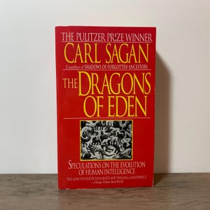 May include: A hardcover book titled "The Dragons of Eden" by Carl Sagan. The book has a red cover with yellow text. The cover includes the text "The Pulitzer Prize Winner" and "Speculations on the Evolution of Human Intelligence".