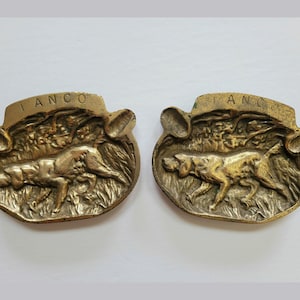 May include: Two brass ashtrays with a hunting dog design. Each ashtray has the word "IANCO" embossed on the top.