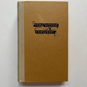 May include: A hardcover book with a tan cover and spine. The title "Vom Winde Verweht" is printed in black on a banner with a sprig of leaves. The spine is a lighter tan color.