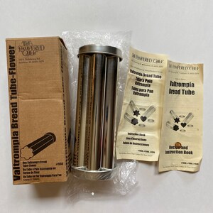 May include: A silver Valtrompia bread tube baking pan with three tubes. The box for the pan is brown and has the text "The Pampered Chef" and "Valtrompia Bread Tube-Flower" on it. The instruction booklet is white with black text and includes the text "Valtrompia Bread Tube" and "Recipe and Instruction Book".