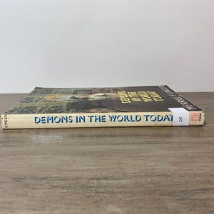 May include: A hardcover book titled "Demons in the World Today" by Merrill F. Unger. The book's spine is light beige with blue text. The cover features a dark image with a person in a white outfit. The book is lying on a wooden surface.