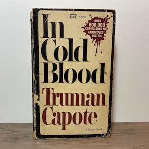 May include: A well-worn paperback copy of "In Cold Blood" by Truman Capote. The cover is a faded tan with black and maroon text. The book has a black border and shows signs of age.